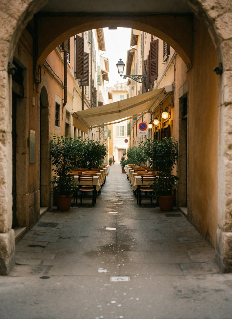 View through a stone archway into a narrow sunlit European lane lined with small outdoor dining tables