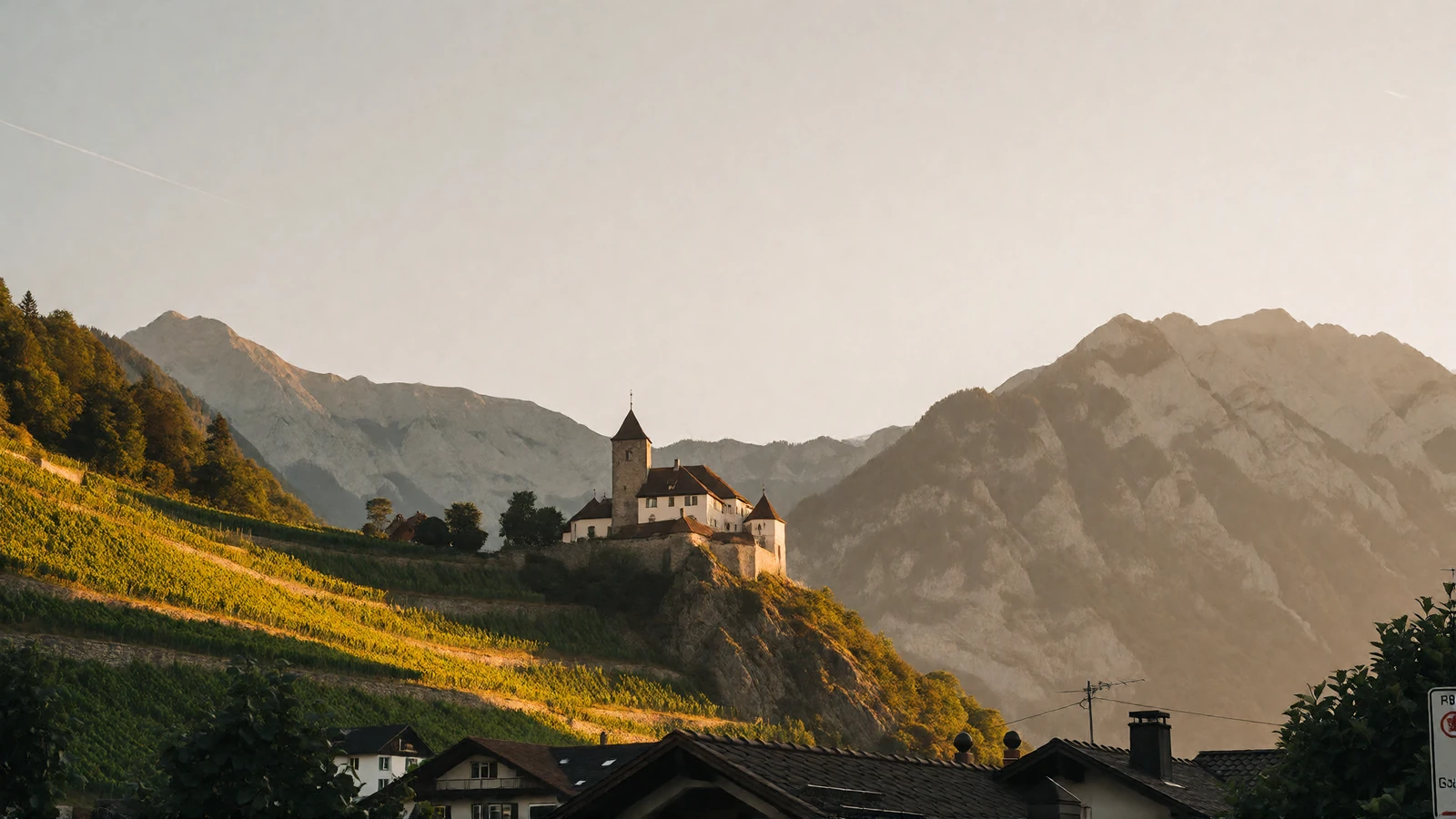 Wide golden-hour view of Vaduz in the Rhine valley with the hillside castle above vineyard terraces and steep alpine ridges behind